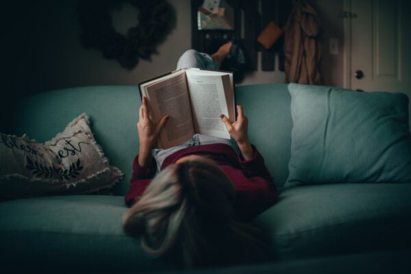 Henkilö makaa sohvalla selällään jalat selkänojalla ja lukee kirjaa.