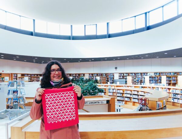Erika poseeraa Kuopion pääkirjaston kirjahallissa iloisenpunaisen Kuopio-kassin kanssa.